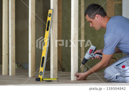 Interior of attic insulated room with oak floor under reconstruction. Young professional worker uses level and screwdriver installing wooden frame for future walls. Renovation and improvement concept. Interior of attic insulated room with oak floor under reconstruction. Young professional worker uses level and screwdriver installing wooden frame for future walls. Renovation and improvement concept. 139304842