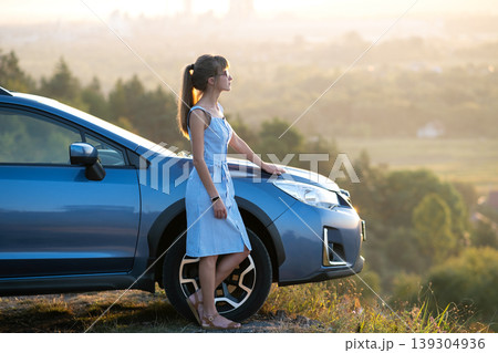 Happy young woman driver in blue dress enjoying warm summer evening standing beside her car. Travelling and vacation concept. Happy young woman driver in blue dress enjoying warm summer evening standing beside her car. Travelling and vacation concept. 139304936