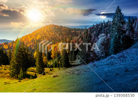 day meet night. fir trees on a meadow down the hill to coniferous forest in mountains of Romania nature. sun and moon over view of remote colorful forest at the foot of a mountain in autumn day meet night. fir trees on a meadow down the hill to coniferous forest in mountains of Romania nature. sun and moon over view of remote colorful forest at the foot of a mountain in autumn 139308530