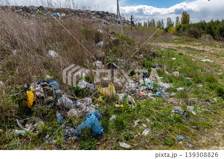 Trash covers green grass in an open space near trees and a distant landfill. This area shows clear signs of pollution and waste Trash covers green grass in an open space near trees and a distant landfill. This area shows clear signs of pollution and waste 139308826