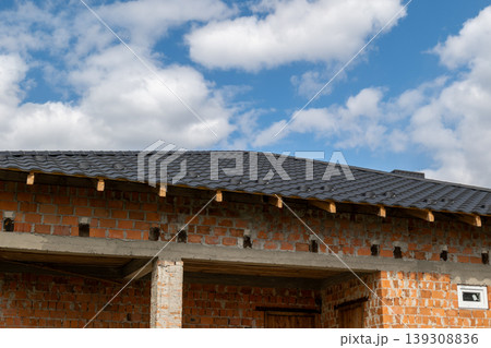 Metal tiles cover the roof of a house under a blue sky with clouds. Construction work is ongoing at the site 139308836