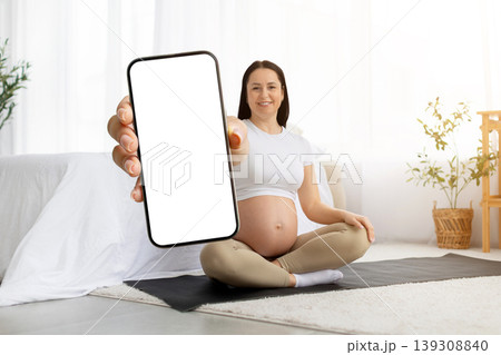 Pregnant young woman sitting on yoga mat holding smartphone with blank screen Pregnant young woman sitting on yoga mat holding smartphone with blank screen 139308840