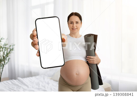 Pregnant woman holding yoga mat under arm and showing blank smartphone screen Pregnant woman holding yoga mat under arm and showing blank smartphone screen 139308841