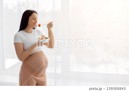 Pregnant woman standing near bright window eating fresh vegetable salad 139308843