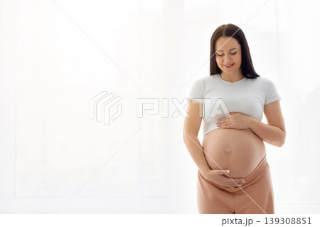 Smiling pregnant woman standing near window tenderly holding belly with both hands 139308851