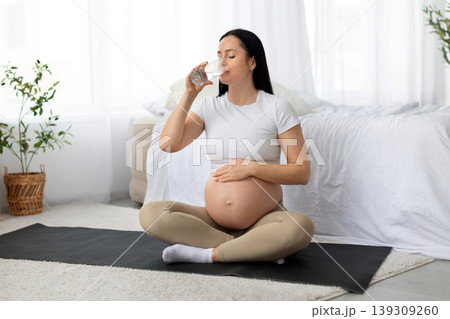 Pregnant young woman drinking water while sitting on yoga mat at home Pregnant young woman drinking water while sitting on yoga mat at home 139309260