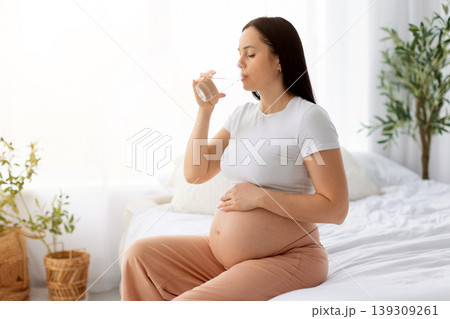 Pregnant woman sitting on bed drinking glass of water while cradling belly Pregnant woman sitting on bed drinking glass of water while cradling belly 139309261