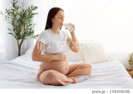 Pregnant woman sitting on bed drinking glass of milk while cradling bare belly Pregnant woman sitting on bed drinking glass of milk while cradling bare belly 139309286