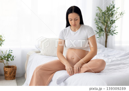 Concerned pregnant woman sitting on bed looking at legs with worried expression Concerned pregnant woman sitting on bed looking at legs with worried expression 139309289