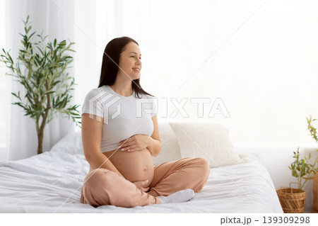 Happy pregnant woman sitting on bed embracing belly, smiling and looking aside 139309298