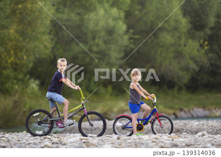 Cute young blond children, boy and girl riding child bicycles on pebbled river bank on blurred green trees bright summer day background. Active lifestyle, recreation and.outdoors activity concept. 139314036