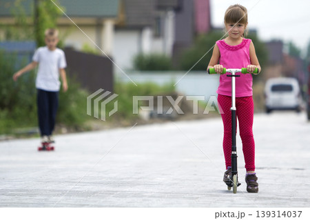 Cute young blond children girl in pink clothing on scooter and handsome boy on skateboard playing on sunny street on blurred summer bright background. Children activities, games and fun concept. 139314037