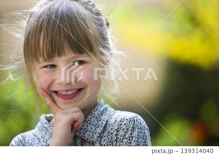 Cute pretty child girl with gray eyes and fair hair smiling in camera outdoors on blurred sunny green and yellow bright bokeh background. Cute pretty child girl with gray eyes and fair hair smiling in camera outdoors on blurred sunny green and yellow bright bokeh background. 139314040