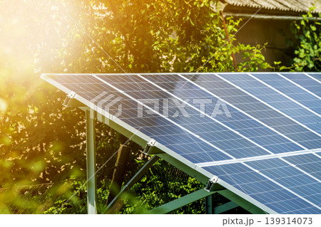 Close-up surface of lit by sun modern saving efficient stand -alone blue shiny solar photo voltaic panels system producing renewable clean energy on green grass and trees foliage background. Close-up surface of lit by sun modern saving efficient stand -alone blue shiny solar photo voltaic panels system producing renewable clean energy on green grass and trees foliage background. 139314073