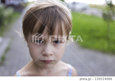 Close-up portrait of pretty young little blond pale unhappy moody friendless child girl looking sadly in camera on blurred sunny outdoors background. Children problems concept. Close-up portrait of pretty young little blond pale unhappy moody friendless child girl looking sadly in camera on blurred sunny outdoors background. Children problems concept. 139314080