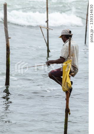 Stilt fisherman with his wooden rod facing side to the camera with his yellow pocket on the pole, fishing in a traditional unique method in Sri Lankan culture, sunny bright evening on the beach. Stilt fisherman with his wooden rod facing side to the camera with his yellow pocket on the pole, fishing in a traditional unique method in Sri Lankan culture, sunny bright evening on the beach. 139314630