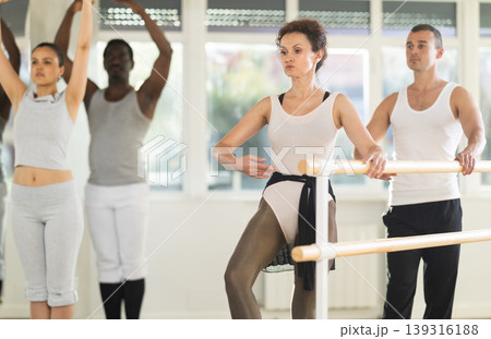 Men and women stand in second position near ballet barre during group training in dance studio Men and women stand in second position near ballet barre during group training in dance studio 139316188