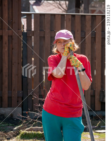Mature woman gardener in red t-shirt leaning on garden tool in backyard. Caucasian lady in baseball cap working outdoors. Agriculture, vegetable garden maintenance and hobby lifestyle concept. 139318219