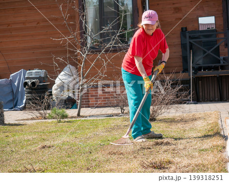 Senior woman gardener raking dry grass in backyard. Elderly lady cleaning lawn with rake in spring. Maintenance of home garden, seasonal outdoor work, healthy active lifestyle for senior. 139318251