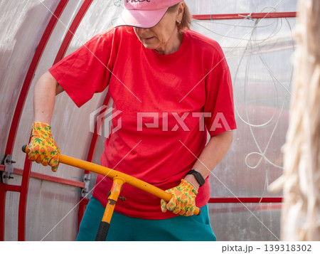 Woman gardener using manual cultivator in greenhouse. Senior lady working with soil tillage tool for agriculture. Cultivation, farming hobby and spring gardening activity concepts. Woman gardener using manual cultivator in greenhouse. Senior lady working with soil tillage tool for agriculture. Cultivation, farming hobby and spring gardening activity concepts. 139318302