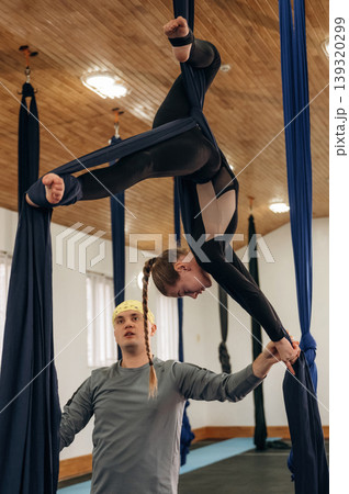 In the air, high up. Male instructor assisting a child in aerial yoga practice on hammock straps in a fitness studio In the air, high up. Male instructor assisting a child in aerial yoga practice on hammock straps in a fitness studio 139320299