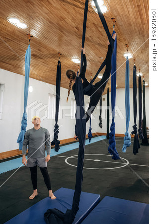 In the air, high up. Male instructor assisting a child in aerial yoga practice on hammock straps in a fitness studio In the air, high up. Male instructor assisting a child in aerial yoga practice on hammock straps in a fitness studio 139320301