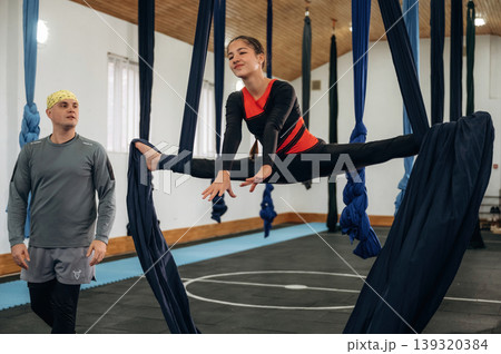 Girl is on hammock straps. Male instructor assisting a child in aerial yoga practice in a fitness studio Girl is on hammock straps. Male instructor assisting a child in aerial yoga practice in a fitness studio 139320384