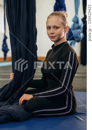 Looking at the camera, sitting on the mat. Child practicing aerial yoga on hammock straps in a fitness studio Looking at the camera, sitting on the mat. Child practicing aerial yoga on hammock straps in a fitness studio 139320425