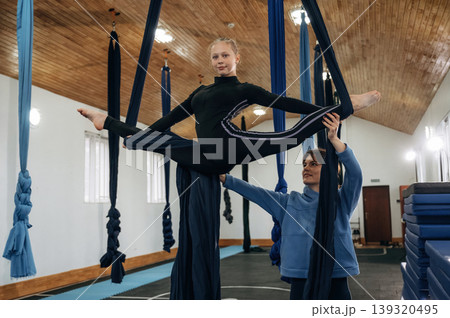 Controlling the twine exercise. Female instructor helping a child practice aerial yoga on hammock straps in a fitness studio 139320495