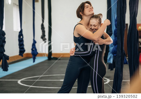 Embracing each other. Female instructor helping a child practice aerial yoga on hammock straps in a fitness studio Embracing each other. Female instructor helping a child practice aerial yoga on hammock straps in a fitness studio 139320502