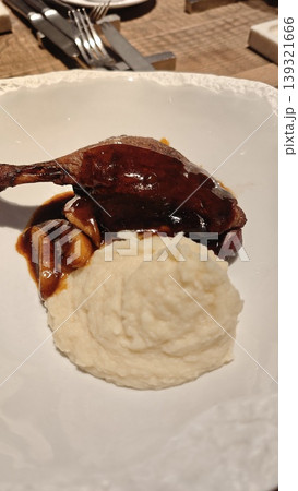 Roasted duck leg accompanied by creamy mashed potatoes and mushroom sauce on a white plate, set on a wooden table with cutlery in the background 139321666