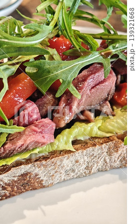Open-faced sandwich featuring tender sliced beef, fresh arugula, and diced tomatoes on whole grain bread, garnished with lettuce and herbs on a white plate Open-faced sandwich featuring tender sliced beef, fresh arugula, and diced tomatoes on whole grain bread, garnished with lettuce and herbs on a white plate 139321668