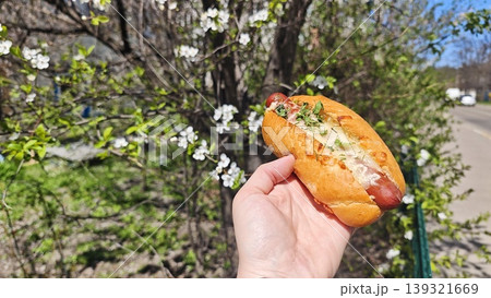 A person holds a hot dog topped with vegetables in front of flowering trees and greenery, capturing a vibrant outdoor scene 139321669