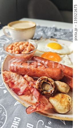 Breakfast plate featuring fried eggs, sausages, bacon, baked beans, and grilled mushrooms served with a cup of coffee on a patterned tablecloth 139321675