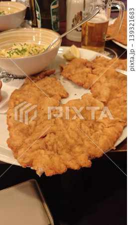 Large breaded schnitzel served on a white plate with a side of pasta salad and a slice of lemon in a restaurant setting with drinks in the background Large breaded schnitzel served on a white plate with a side of pasta salad and a slice of lemon in a restaurant setting with drinks in the background 139321683