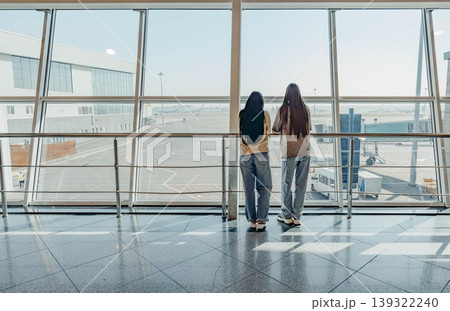 Two young women, one with long black hair and the other with long brown hair, stand by large window at an airport, looking out at the runway Two young women, one with long black hair and the other with long brown hair, stand by large window at an airport, looking out at the runway 139322240