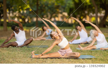 Group of people of multinational practicing hatha yoga in summer garden, stretching in an asymmetrical seated asana Parivritta Sirsasana 139324902