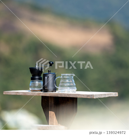 The coffee grinder, kettle, and coffee pot arranged on the table at the beautiful flower field view above the forest and mountains background The coffee grinder, kettle, and coffee pot arranged on the table at the beautiful flower field view above the forest and mountains background 139324972