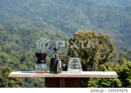The coffee grinder, kettle, and coffee pot arranged on the table at the beautiful flower field view above the forest and mountains background The coffee grinder, kettle, and coffee pot arranged on the table at the beautiful flower field view above the forest and mountains background 139324976