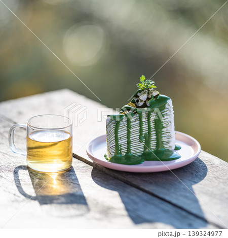The shot of a hot Oolong tea cup and delicious matcha green tea crepe cake dish placed on the wooden table under the sunlight over the blurry background. The shot of a hot Oolong tea cup and delicious matcha green tea crepe cake dish placed on the wooden table under the sunlight over the blurry background. 139324977