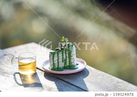 The shot of a hot Oolong tea cup and delicious matcha green tea crepe cake dish placed on the wooden table under the sunlight over the blurry background. The shot of a hot Oolong tea cup and delicious matcha green tea crepe cake dish placed on the wooden table under the sunlight over the blurry background. 139324978