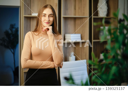 Woman stands in office space smiling while resting her chin on her hand in natural light 139325137
