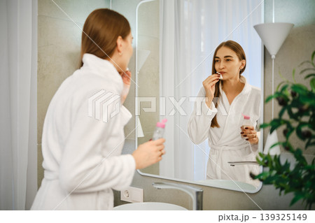 Woman in bathroom preparing for her day while looking in the mirror and applying skincare Woman in bathroom preparing for her day while looking in the mirror and applying skincare 139325149