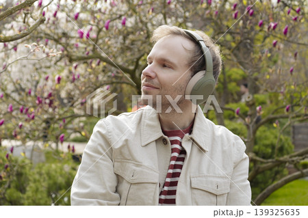 A smiling man with headphones relaxes in nature among blooming magnolias in a Japanese park, enjoying a digital detox. A smiling man with headphones relaxes in nature among blooming magnolias in a Japanese park, enjoying a digital detox. 139325635