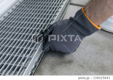 A worker wearing safety gloves removes a metal drain grate, illustrating a hands on drainage maintenance process A worker wearing safety gloves removes a metal drain grate, illustrating a hands on drainage maintenance process 139325641