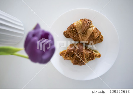 Tho croissants on a plate and purple tulip in a vase in the foreground. Tho croissants on a plate and purple tulip in a vase in the foreground. 139325658