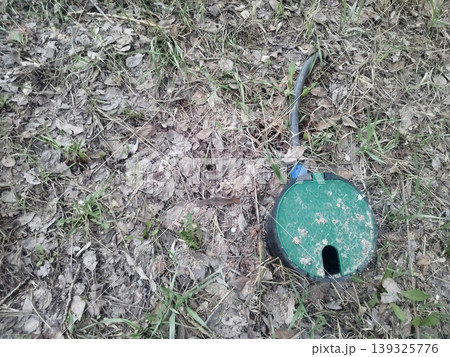 Green plastic irrigation valve box lid sits on dry ground covered in dead leaves and sparse grass. A black pipe extends from the circular box, indicating an underground garden watering system Green plastic irrigation valve box lid sits on dry ground covered in dead leaves and sparse grass. A black pipe extends from the circular box, indicating an underground garden watering system 139325776