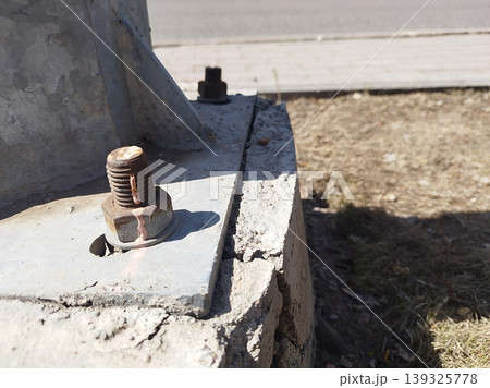 Close-up shows a large, rusted metal bolt and nut securing the base of a structural pole to a cracked concrete foundation. The worn materials and damaged cement highlight urban infrastructure decay Close-up shows a large, rusted metal bolt and nut securing the base of a structural pole to a cracked concrete foundation. The worn materials and damaged cement highlight urban infrastructure decay 139325778