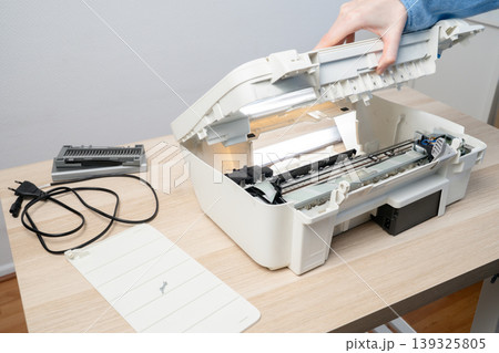 Person performing maintenance on an open white inkjet printer scanner on desk, showing internal components, power cable and removed parts, illustrating repair, troubleshooting and office technology. Person performing maintenance on an open white inkjet printer scanner on desk, showing internal components, power cable and removed parts, illustrating repair, troubleshooting and office technology. 139325805