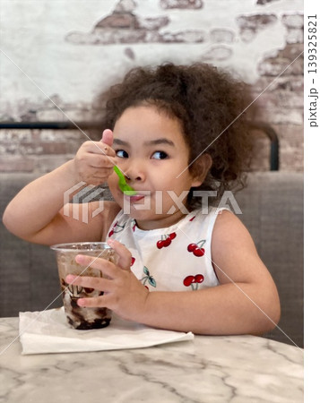 Portrait of cute Asian little girl with curly hair eating chocolate ice cream in a glass with a green spoon in her hand at a cafe 139325821